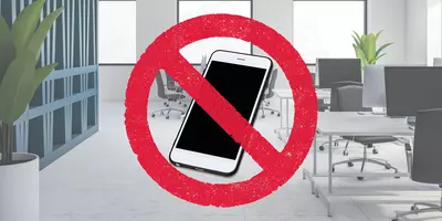 Das Bild zeigt ein modernes Büro mit einem durchgestrichenen Smartphone in einem roten Verbotssymbol, das auf ein Handyverbot hinweist. Im Hintergrund sind Schreibtische, Stühle und Pflanzen zu sehen.