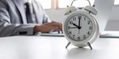 Das Bild zeigt einen Schreibtisch mit einer Uhr im Vordergrund und einem Menschen im Anzug im Hintergrund. Die Uhr steht symbolisch für das Erfassen der Arbeitszeit.