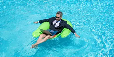 Das Bild zeigt einen Mann auf einer Luftmatratze in einem Schwimmbecken. Er trägt ein Jackett und hat einen Laptop auf dem Schoß, was symbolisch für Homeoffice im Freien steht.