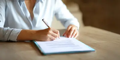 Auf dem Bild ist der Oberkörper einer Person zu sehen, die an einem Schreibtisch vor einem Blatt Papier sitzt. Das Papier stellt ein Arbeitszeugnis dar.