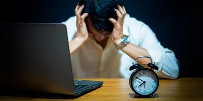 Ein Mann sitzt vor einem Laptop und stützt den Kopf in die Hände, was Frustration, Erschöpfung oder Kopfschmerzen darstellt. Die Uhr vor ihm zeigt, dass er Überstunden macht.