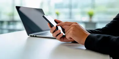 Das Bild zeigt eine Person an einem Schreibtisch. In der Hand hält sie ein Handy, im Hintergrund ist ein Laptop zu sehen. Bei diesen elektronischen Geräten handelt es sich um Arbeitsgeräte.