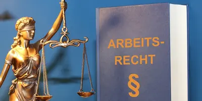 Das Bild zeigt ein blaues Buch mit der Aufschrift "Arbeitsrecht". Daneben ist eine goldene Justizia-Figur zu sehen.