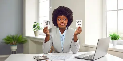 Eine Frau sitzt im Büro am Schreibtisch. Sie hat einen ratlosen Gesichtsausdruck und hält zwei Karten mit den Aufschriften "Yes" und "No" in der Hand. Dies steht symbolisch für die Überlegung, ob es sinnvoll ist, ohne neuen Job zu kündigen.